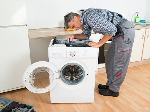 Full length of handyman checking washing machine with flashlight in kitchen